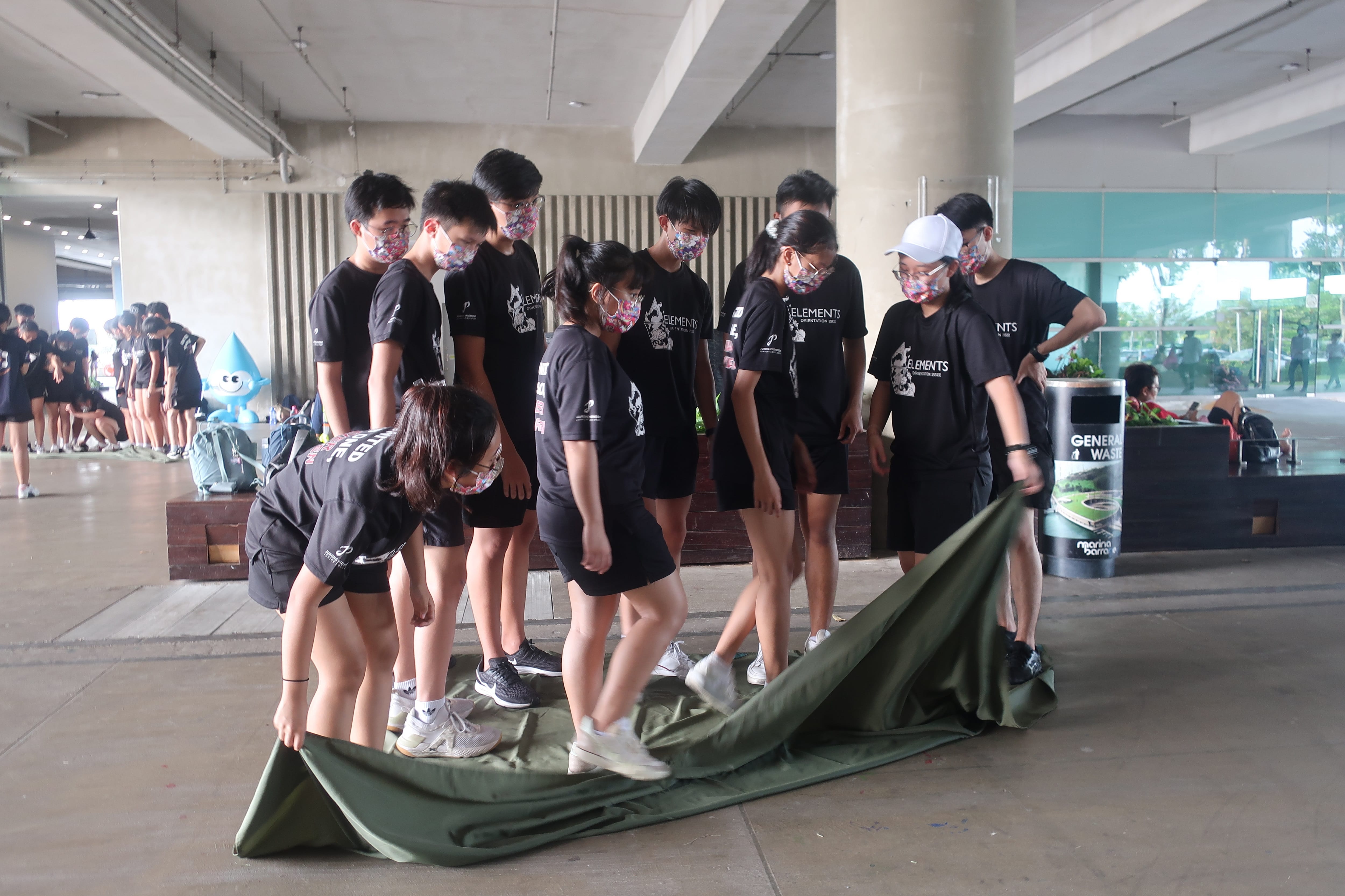 A group of masked teens in black shirts spread a large green tarp on the ground under a covered outdoor area.